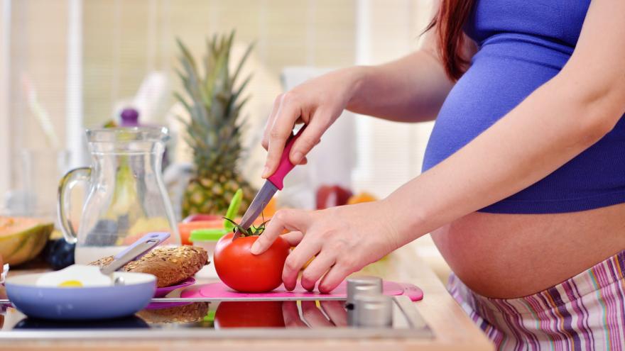 Las manos de una mujer embarazada cortan un tomate sobre la encimera de la cocina